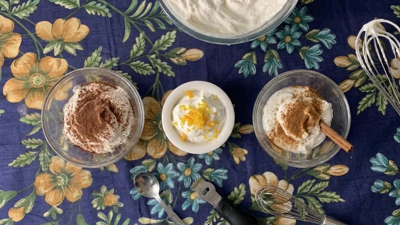 Several small bowls of whipped cream on a counter, one with cocoa powder, one with citrus zest, and one with cinnamon nearby
