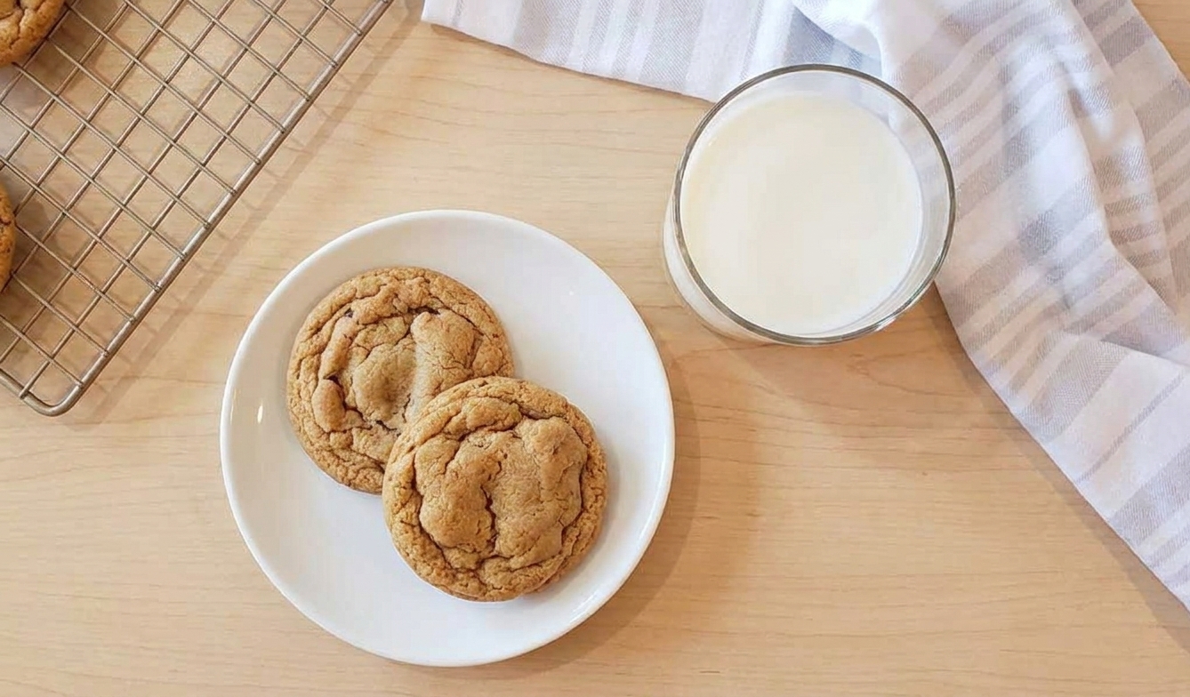 Two brown sugar cookies on a small plate next to a glass of milk on a wooden table